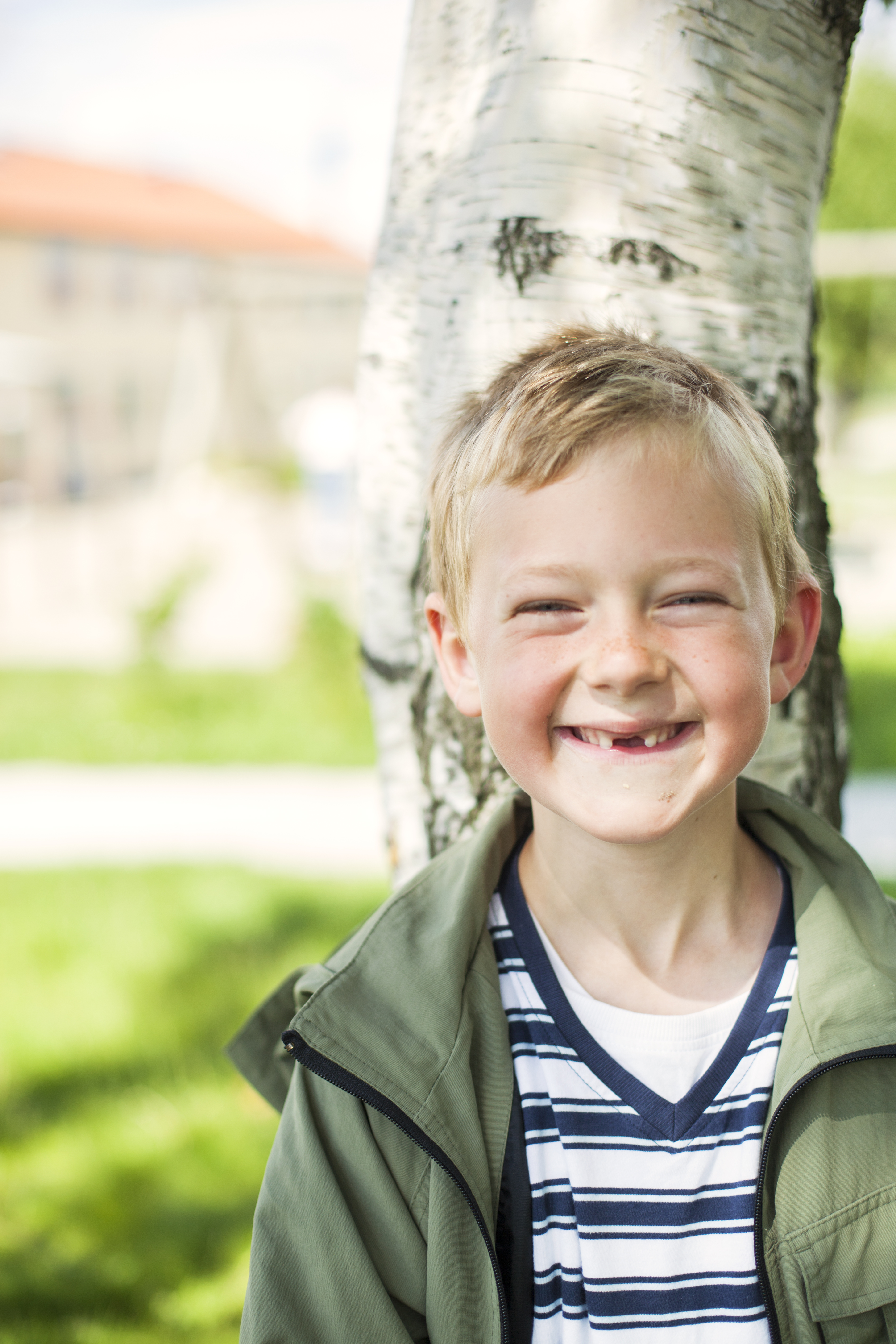 Portrait of happy schoolboy leaning on tree trunk at yard Smiling young boy standing outdoors in front of a tree and wearing a green jacket over a striped shirt